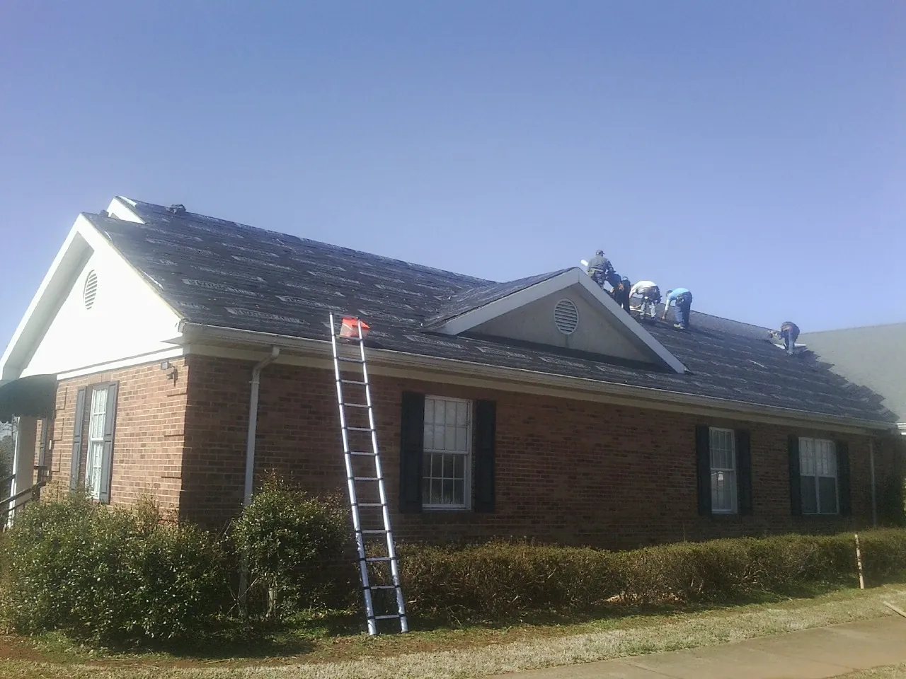 Crew tearing shingles off a brick ranch roof with a ladder set — Emergency Roof Repair work by our crew in Rock Valley