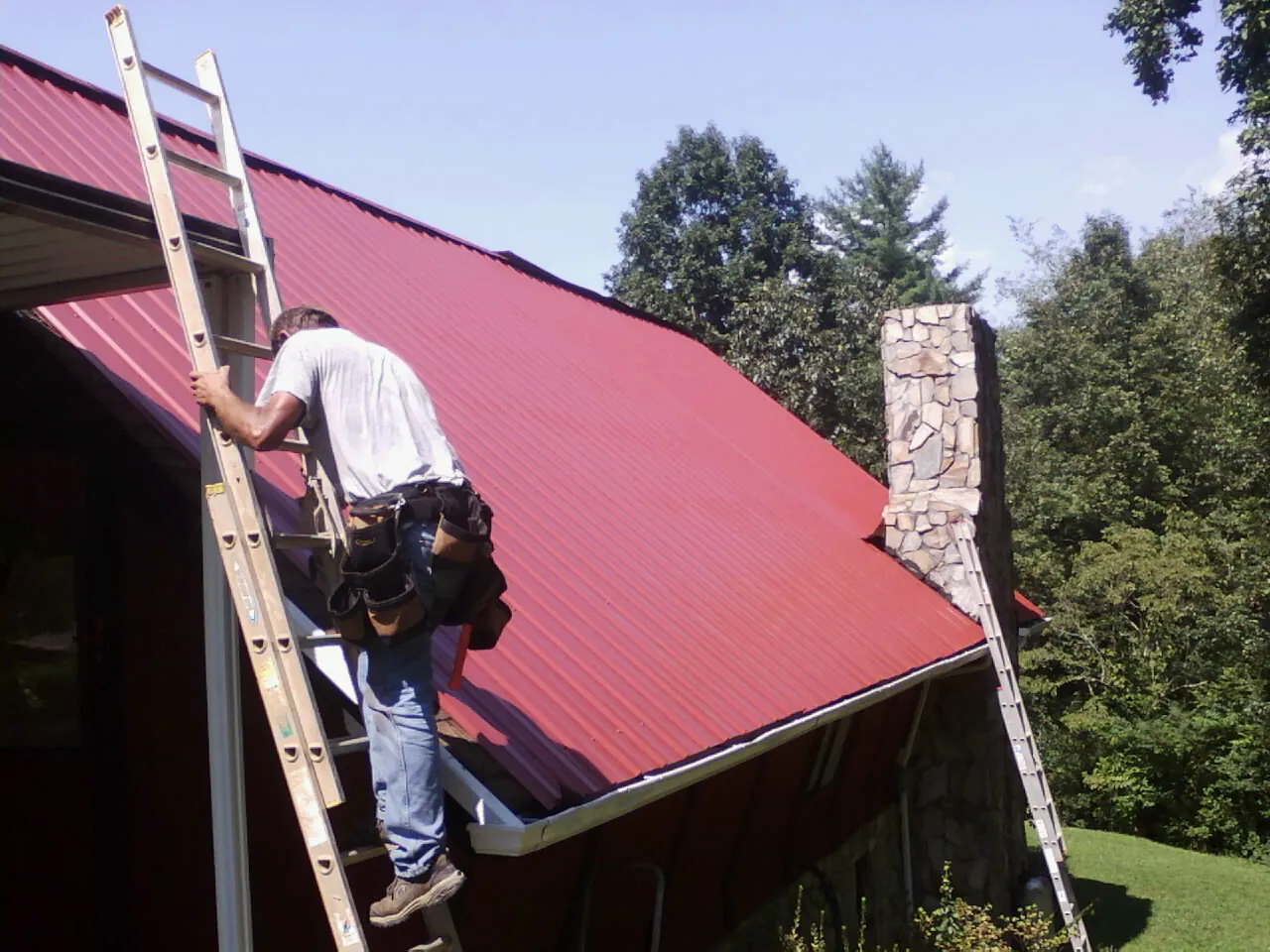 Roofer on a ladder beside a red metal roof and stone chimney — Soffit & Fascia Repair by Roofing Brothers in Rock Valley